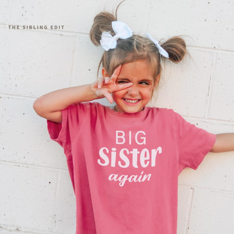 Puede incluir: Una ni&ntilde;a sonr&iacute;e, vistiendo una camiseta rosa con las palabras "BIG Sister again" en blanco. Tiene dos mo&ntilde;os en el pelo, cada uno adornado con un lazo blanco. El fondo es una pared de ladrillos blancos. THE SIBLING EDIT est&aacute; en la esquina superior izquierda.