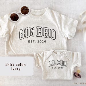 Puede incluir: Camisetas color marfil, una para adulto y otra para beb&eacute;, con texto negro. La camiseta de adulto dice "BIG BRO EST. 2026", mientras que la camiseta de beb&eacute; dice "LIL BRO EST. 2026". Gafas de sol blancas sobre la camiseta de adulto.