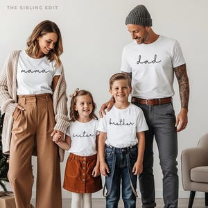 May include: A family of five wearing matching white t-shirts with black script. The shirts read "mama", "dad", "sister", and "brother". The adults wear neutral-colored pants and a cardigan. The children wear a corduroy skirt and jeans.