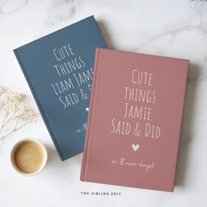 May include: Two hardcover journals, one dusty blue and one rose-colored, with white text reading "CUTE THINGS [NAME] SAID & DID." A small heart graphic is below the text. A cup of coffee sits nearby.