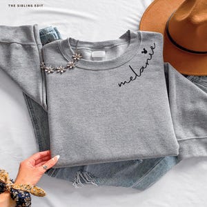 May include: A grey sweatshirt with the name "melanie" in cursive script. A small Mickey Mouse head is next to the name. A floral necklace and a brown hat are also in the image.