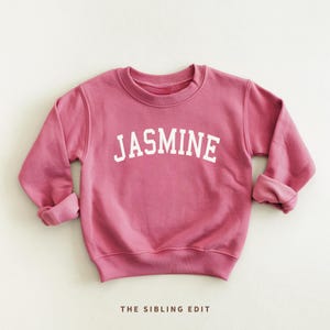 May include: A dusty rose-colored sweatshirt with the name "JASMINE" printed in white block letters across the chest. The sleeves are cuffed. The text "THE SIBLING EDIT" is printed in small white letters at the bottom.