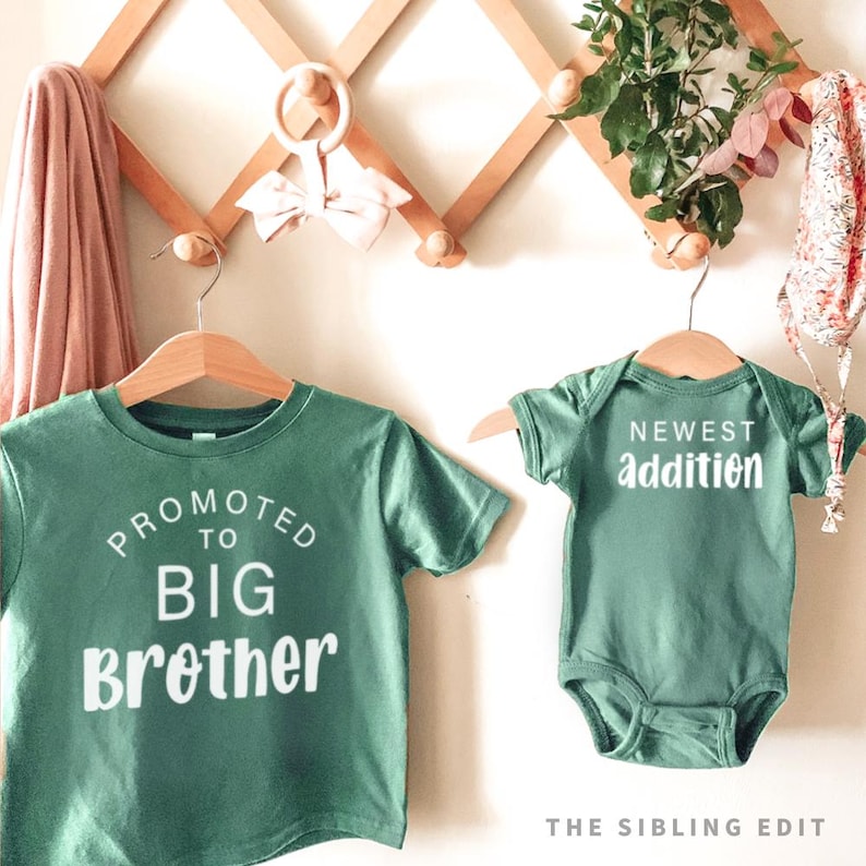 Puede incluir: Dos camisetas verdes en perchas de madera. Una dice "PROMOTED TO BIG Brother" en texto blanco. La otra es un body de beb&eacute; con "NEWEST addition" en texto blanco. Las camisas est&aacute;n colgadas en un estante de pared de madera con elementos decorativos.
