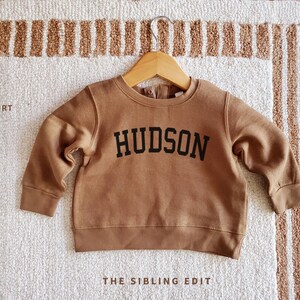 May include: A brown infant sweatshirt with the name "HUDSON" in black block letters. The sweatshirt is hanging on a wooden hanger against a neutral-toned rug. The text "INFANT SWEATSHIRT" and "THE SIBLING EDIT" are also visible.