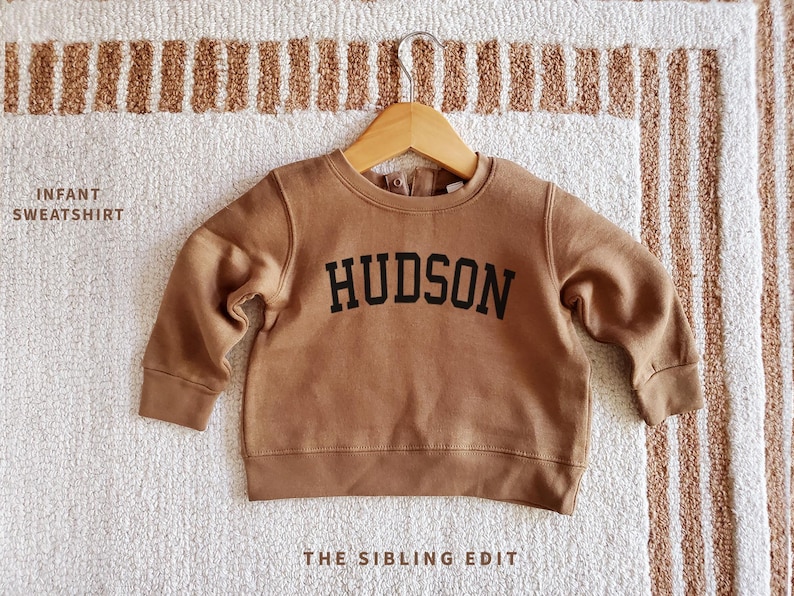 May include: A brown infant sweatshirt with the name "HUDSON" in black block letters. The sweatshirt is hanging on a wooden hanger against a neutral-toned rug. The text "INFANT SWEATSHIRT" and "THE SIBLING EDIT" are also visible.