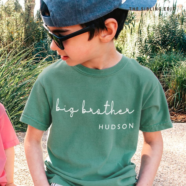 May include: A green t-shirt with the words "big brother" in white script, with the name "HUDSON" below. The shirt is worn by a person wearing sunglasses and a blue cap.