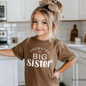 Puede incluir: Una ni&ntilde;a peque&ntilde;a con una camiseta marr&oacute;n con el texto blanco "PROMOTED TO BIG SISTER". Tiene el pelo rubio y un lazo a juego. El fondo es una cocina con armarios y electrodom&eacute;sticos blancos.