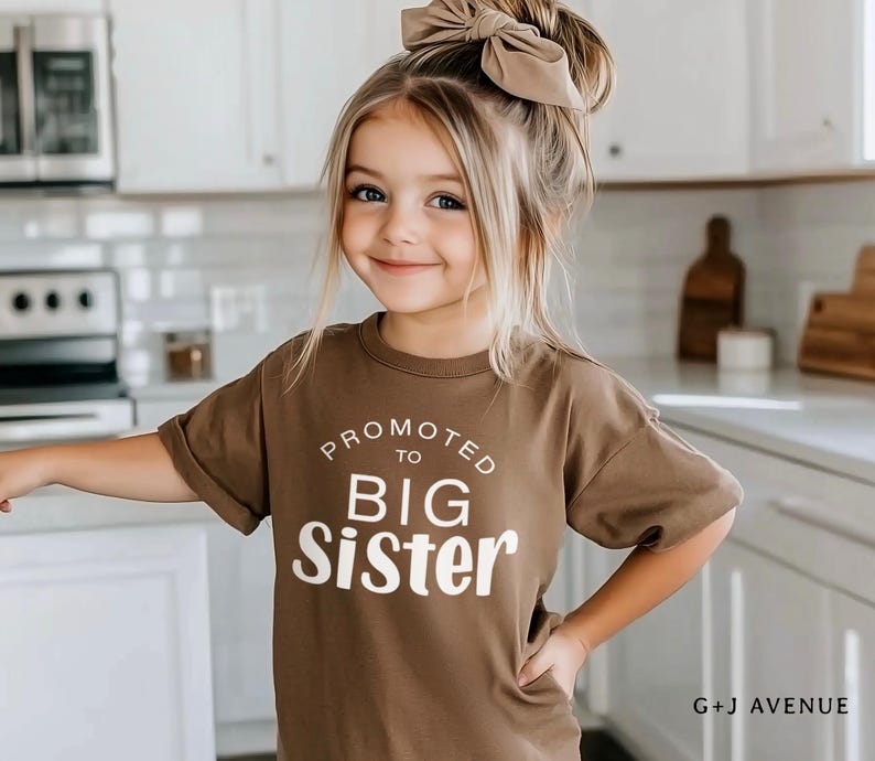 Puede incluir: Una ni&ntilde;a peque&ntilde;a con una camiseta marr&oacute;n con el texto blanco "PROMOTED TO BIG SISTER". Tiene el pelo rubio y un lazo a juego. El fondo es una cocina con armarios y electrodom&eacute;sticos blancos.