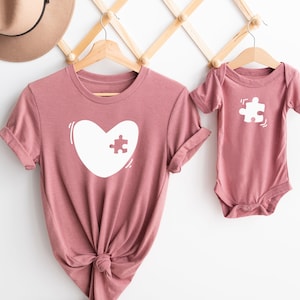 May include: Two shirts with a puzzle piece design. The larger shirt is a dusty rose color with a white heart shape with a puzzle piece missing. The smaller shirt is a dusty rose bodysuit with a white puzzle piece.
