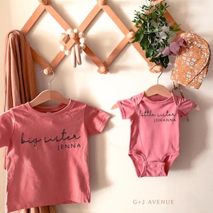 May include: Two pink shirts, one a t-shirt and the other a onesie, hang on wooden hangers. The t-shirt reads "big sister" and the onesie says "little sister". The text "G + J AVENUE" is at the bottom.