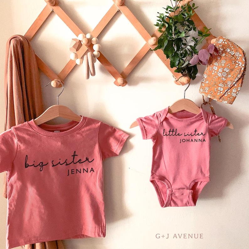 May include: Two pink shirts, one a t-shirt and the other a onesie, hang on wooden hangers. The t-shirt reads "big sister" and the onesie says "little sister". The text "G + J AVENUE" is at the bottom.