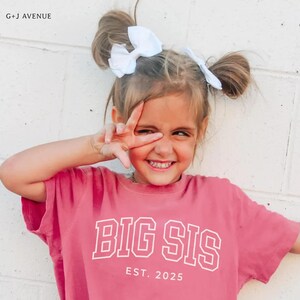Puede incluir: Una ni&ntilde;a sonr&iacute;e, vistiendo una camiseta rosa con las palabras "BIG SIS" y "EST. 2025" en letras blancas. Tiene dos lazos blancos en el pelo. La imagen est&aacute; tomada contra una pared de ladrillos blancos.