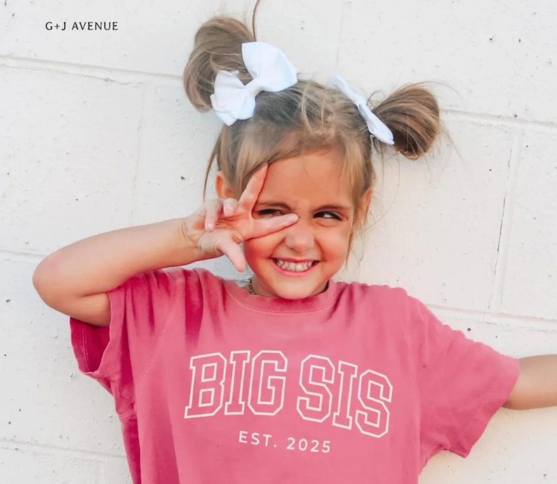 Puede incluir: Una ni&ntilde;a sonr&iacute;e, vistiendo una camiseta rosa con las palabras "BIG SIS" y "EST. 2025" en letras blancas. Tiene dos lazos blancos en el pelo. La imagen est&aacute; tomada contra una pared de ladrillos blancos.