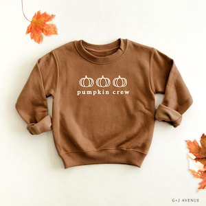 May include: A brown long-sleeve crewneck sweatshirt featuring a white graphic of three pumpkins arranged horizontally above the text "pumpkin crew" in a clean, sans-serif font. The garment has ribbed cuffs and a ribbed neckline.