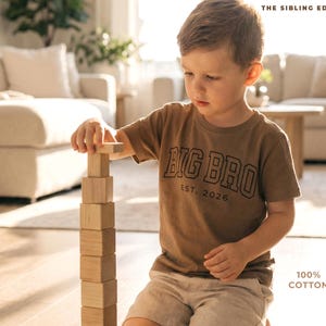 Puede incluir: Un ni&ntilde;o con una camiseta marr&oacute;n con la frase "BIG BRO EST. 2026" construye una torre con bloques de madera. La camiseta es 100% algod&oacute;n. La imagen est&aacute; ambientada en una habitaci&oacute;n luminosa con tonos neutros.