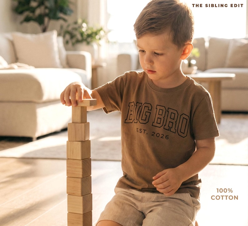Puede incluir: Un ni&ntilde;o con una camiseta marr&oacute;n con la frase "BIG BRO EST. 2026" construye una torre con bloques de madera. La camiseta es 100% algod&oacute;n. La imagen est&aacute; ambientada en una habitaci&oacute;n luminosa con tonos neutros.