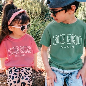Puede incluir: Dos ni&ntilde;os con gafas de sol y camisetas a juego. La camiseta de la ni&ntilde;a es rosa y dice "BIG SIS EST. 2026". La camiseta del ni&ntilde;o es verde y dice "BIG BRO AGAIN". Est&aacute;n al aire libre.