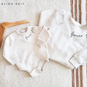 May include: Two matching light beige sweatshirts, one a baby onesie, the other a crew neck. Both have the words "Flower girl" in black script. A wooden hanger is in the upper left corner.