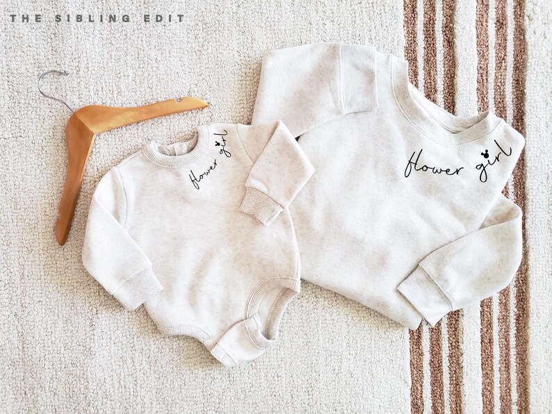 May include: Two matching light beige sweatshirts, one a baby onesie, the other a crew neck. Both have the words "Flower girl" in black script. A wooden hanger is in the upper left corner.
