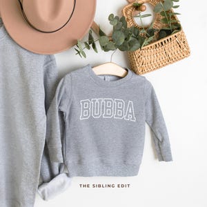 May include: A gray sweatshirt with the word "BUBBA" in white, collegiate-style lettering. The sweatshirt is hanging on a wooden hanger. A matching gray sweatshirt and a brown hat are visible in the background. The text "THE SIBLING EDIT" is at the bottom.