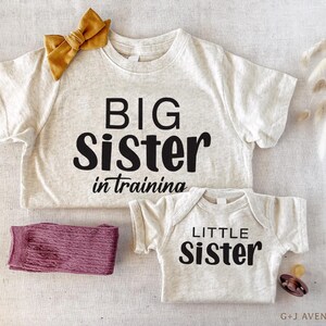Puede incluir: Una camiseta de color beige claro con las palabras "BIG Sister in training" en letras negras, combinada con un body a juego que dice "LITTLE Sister". Un lazo amarillo mostaza y un par de calcetines malva tambi&eacute;n est&aacute;n en la imagen.
