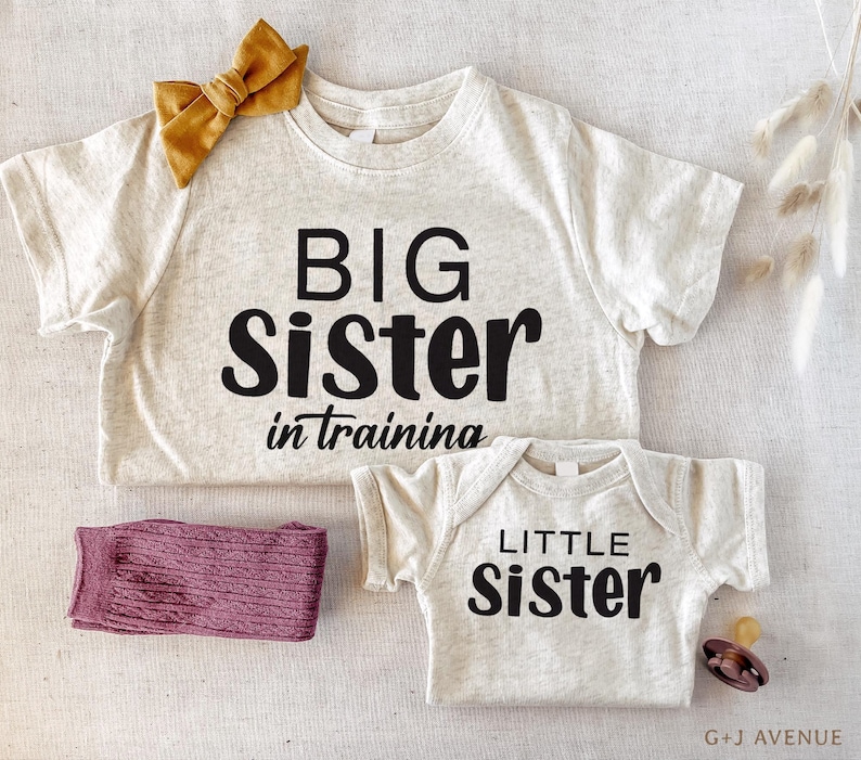 Puede incluir: Una camiseta de color beige claro con las palabras "BIG Sister in training" en letras negras, combinada con un body a juego que dice "LITTLE Sister". Un lazo amarillo mostaza y un par de calcetines malva tambi&eacute;n est&aacute;n en la imagen.