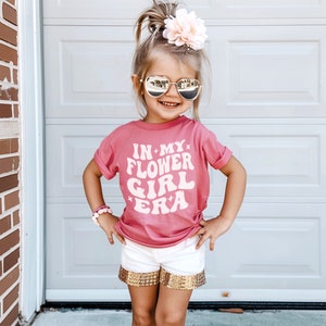 May include: A young girl wearing a pink t-shirt with the text "In My Flower Girl Era" in white. She is wearing white shorts with gold sequins and has a pink flower in her hair.