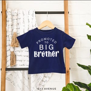 Puede incluir: Una camiseta azul marino de manga corta con el texto blanco "PROMOTED TO BIG Brother". La camiseta est&aacute; colgada de una percha de madera. Una manta blanca con las palabras "I love you" est&aacute; en el fondo.