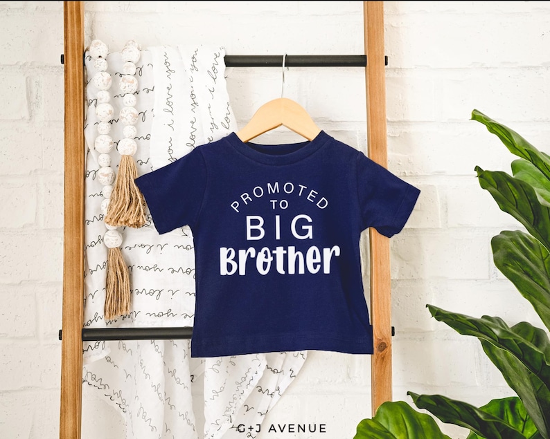 Puede incluir: Una camiseta azul marino de manga corta con el texto blanco "PROMOTED TO BIG Brother". La camiseta est&aacute; colgada de una percha de madera. Una manta blanca con las palabras "I love you" est&aacute; en el fondo.