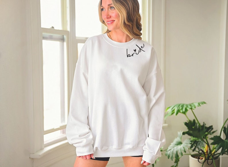 May include: White sweatshirt with the word "bride" in black script on the upper left chest. The word "bride" has two small hearts above the "i". The sweatshirt has long sleeves and a crew neck.