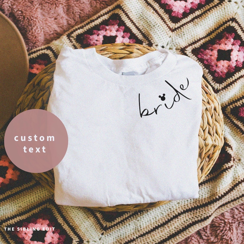 May include: A white t-shirt with the word "bride" in black script, with a Mickey Mouse head as the dot of the "i". The shirt is folded on a woven basket, with a custom text label and the text "THE SIBLING EDIT".