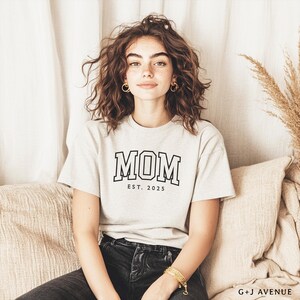 Puede incluir: Camiseta gris claro con la palabra "MOM" en letras negras y "EST. 2025" debajo. La camiseta la lleva una persona sentada en un coj&iacute;n beige.