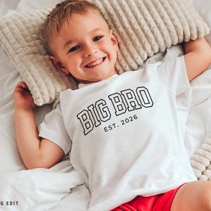 Puede incluir: Un ni&ntilde;o peque&ntilde;o con una camiseta blanca con el texto "BIG BRO EST. 2026" en negro. El ni&ntilde;o sonr&iacute;e y est&aacute; acostado en una cama con una almohada beige. El ni&ntilde;o lleva pantalones cortos rojos.