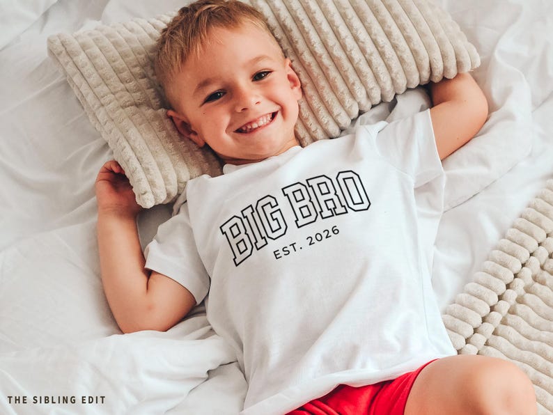 Puede incluir: Un ni&ntilde;o peque&ntilde;o con una camiseta blanca con el texto "BIG BRO EST. 2026" en negro. El ni&ntilde;o sonr&iacute;e y est&aacute; acostado en una cama con una almohada beige. El ni&ntilde;o lleva pantalones cortos rojos.