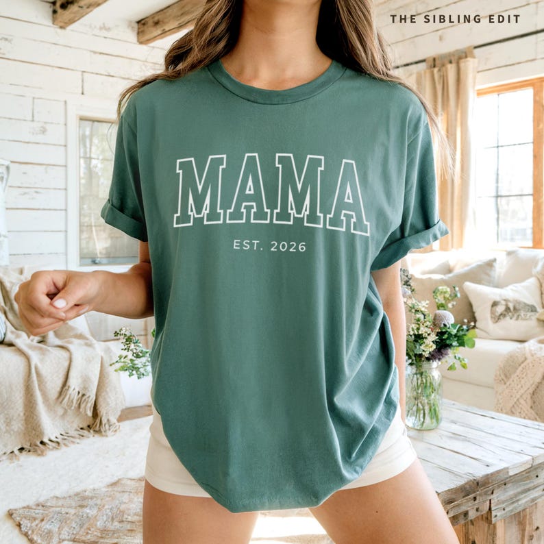Puede incluir: Camiseta verde con la palabra "MAMA" en letras blancas con contorno blanco. Debajo de la palabra "MAMA" est&aacute; el texto "EST. 2026". La camiseta es de cuello redondo.