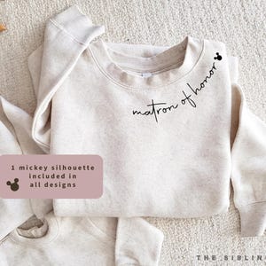 May include: A light beige sweatshirt with the words "matron of honor" in black script. A small Mickey Mouse silhouette is above the text. The sweatshirt is folded on a light-coloured surface. The text "1 mickey silhouette included in all designs" is in a pink rectangle.