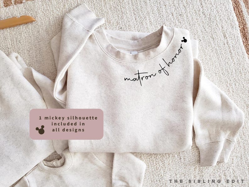 May include: A light beige sweatshirt with the words "matron of honor" in black script. A small Mickey Mouse silhouette is above the text. The sweatshirt is folded on a light-coloured surface. The text "1 mickey silhouette included in all designs" is in a pink rectangle.