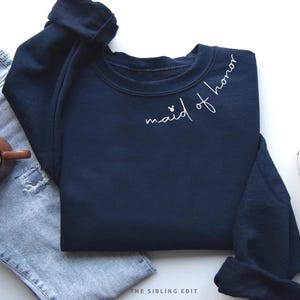 May include: A navy blue sweatshirt with the words "maid of honor" in white script. The sweatshirt is paired with light blue jeans, a brown mug of coffee, and a grey trainer. The Sibling Edit logo is visible.