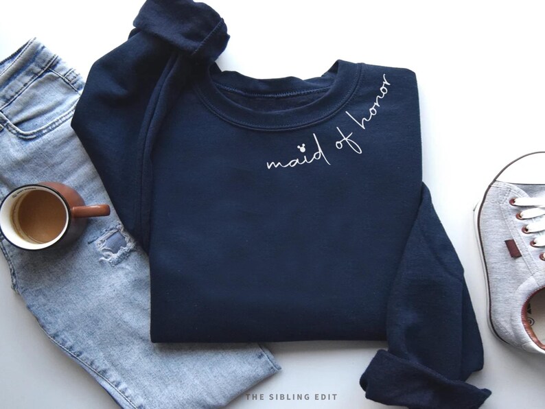 May include: A navy blue sweatshirt with the words "maid of honor" in white script. The sweatshirt is paired with light blue jeans, a brown mug of coffee, and a grey trainer. The Sibling Edit logo is visible.