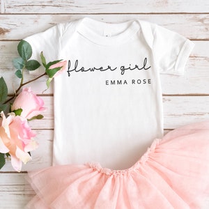 May include: White baby bodysuit with the text "flower girl" and the name "EMMA ROSE" printed in black. The bodysuit is paired with a pink tulle tutu.