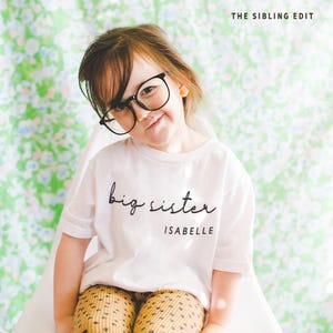 May include: A young child wearing a white t-shirt with the words "big sister" and the name "Isabelle" in black script. The child is also wearing black-framed glasses and patterned leggings. The background features a floral pattern, and the text "THE SIBLING EDIT" is visible.