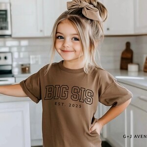 Puede incluir: Una ni&ntilde;a peque&ntilde;a con una camiseta marr&oacute;n con el texto "BIG SIS EST. 2025" en negro. Tiene el pelo rubio y un lazo grande. El fondo es una cocina con armarios y electrodom&eacute;sticos blancos.