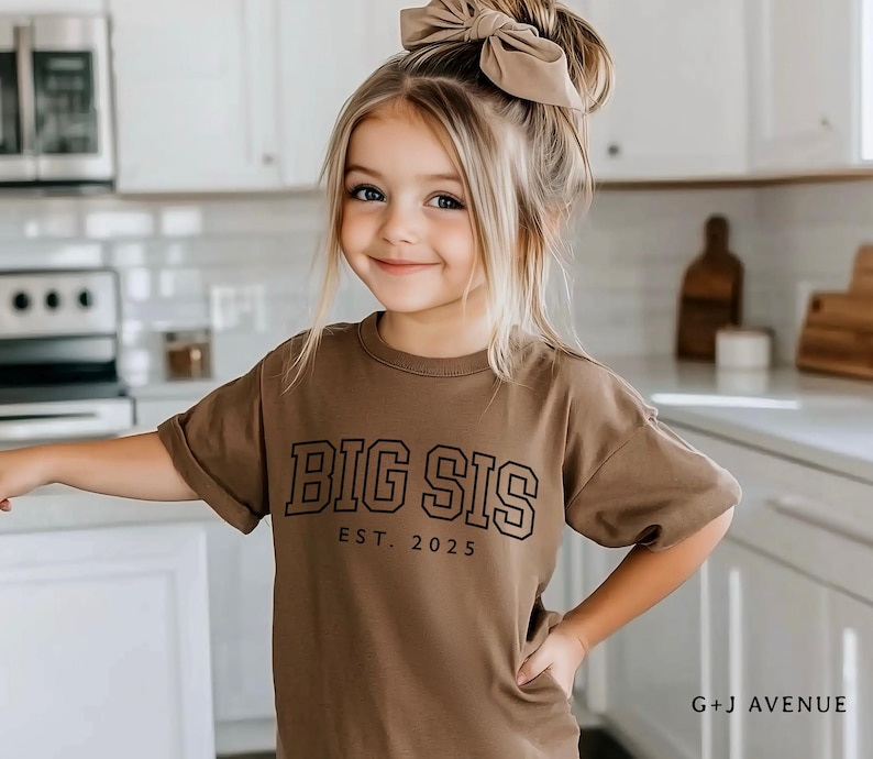 Puede incluir: Una ni&ntilde;a peque&ntilde;a con una camiseta marr&oacute;n con el texto "BIG SIS EST. 2025" en negro. Tiene el pelo rubio y un lazo grande. El fondo es una cocina con armarios y electrodom&eacute;sticos blancos.