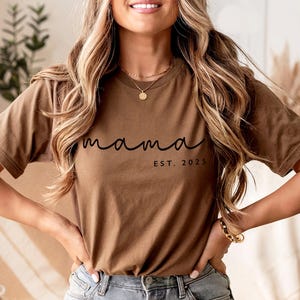May include: A brown t-shirt with the word "mama" in a cursive font and the text "EST. 2025" in a smaller, block font. The shirt is made of a soft material and has short sleeves. The model is wearing the shirt with blue jeans.