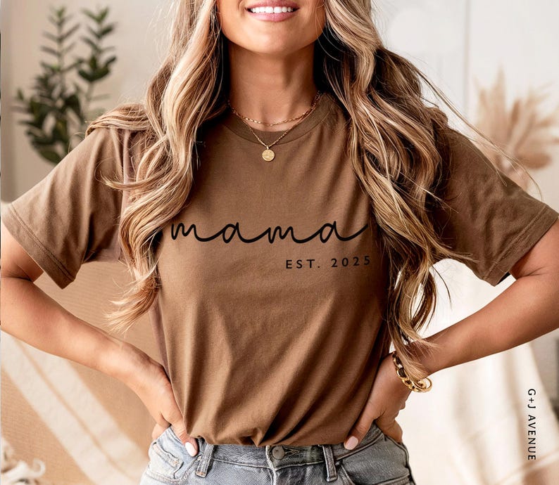 May include: A brown t-shirt with the word "mama" in a cursive font and the text "EST. 2025" in a smaller, block font. The shirt is made of a soft material and has short sleeves. The model is wearing the shirt with blue jeans.