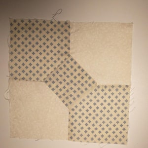May include: A square quilt block composed of four fabric pieces. Two cream-colored squares are paired with two patterned pieces featuring a light yellow background and a repeating blue floral design. The pieces are sewn together.