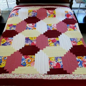 May include: A quilt with a log cabin pattern in shades of pink, burgundy, white, and yellow. The quilt features floral fabric with a variety of colors and patterns.