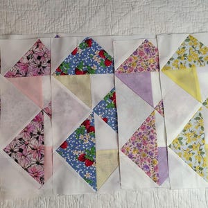May include: A collection of quilt blocks with various floral patterns and solid colors. The blocks feature triangles of yellow, purple, pink, and blue floral fabrics, and white fabric. The blocks are arranged in a row.