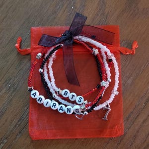 Könnte beinhalten: Ein Set aus Perlenarmbändern in Rot, Schwarz und Weiß, mit silbernen Akzenten und Buchstabenperlen mit der Aufschrift "ARIRANG" und "BTS". Die Armbänder werden in einem roten Kordelzugbeutel mit schwarzem Band präsentiert.