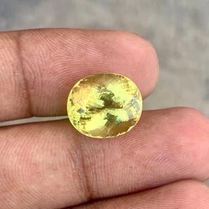 May include: A faceted, oval-shaped gemstone with a pale yellow hue. The gem is held between fingers, showcasing its polished surface and internal light reflections. The gemstone's cut reveals multiple facets, enhancing its brilliance.